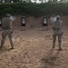 Two military personnel in full combat gear and camouflage uniforms are conducting pistol qualification or training at an outdoor shooting range. The left shooter is actively firing, as evidenced by visible muzzle smoke, while the right shooter stands in a ready position. Four standard silhouette targets are positioned downrange against a sandy berm backdrop.