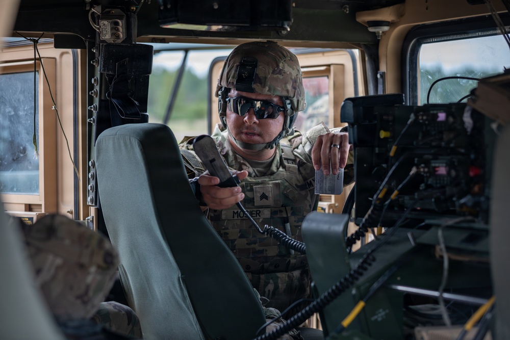 A U.S. Army soldier, identified by name tape as 'MOSIER' with Sergeant rank insignia, is operating communications equipment inside a military HMMWV (Humvee). The soldier is wearing full combat gear including an OCP (Operational Camouflage Pattern) uniform, body armor, ballistic helmet, and tactical sunglasses. They appear to be referencing a small card while handling a radio handset, suggesting active radio communication or equipment configuration.