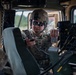 A U.S. Army soldier, identified by name tape as 'MOSIER' with Sergeant rank insignia, is operating communications equipment inside a military HMMWV (Humvee). The soldier is wearing full combat gear including an OCP (Operational Camouflage Pattern) uniform, body armor, ballistic helmet, and tactical sunglasses. They appear to be referencing a small card while handling a radio handset, suggesting active radio communication or equipment configuration.