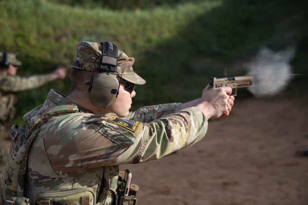 A soldier in MultiCam/OCP camouflage uniform is actively firing a pistol at a shooting range, with visible muzzle blast/gas discharge captured at the moment of firing. The shooter is wearing hearing protection earmuffs, sunglasses, a patrol cap with a US flag patch, and body armor with tactical pouches. At least one additional uniformed personnel is visible in the blurred background.