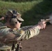 A soldier in MultiCam/OCP camouflage uniform is actively firing a pistol at a shooting range, with visible muzzle blast/gas discharge captured at the moment of firing. The shooter is wearing hearing protection earmuffs, sunglasses, a patrol cap with a US flag patch, and body armor with tactical pouches. At least one additional uniformed personnel is visible in the blurred background.