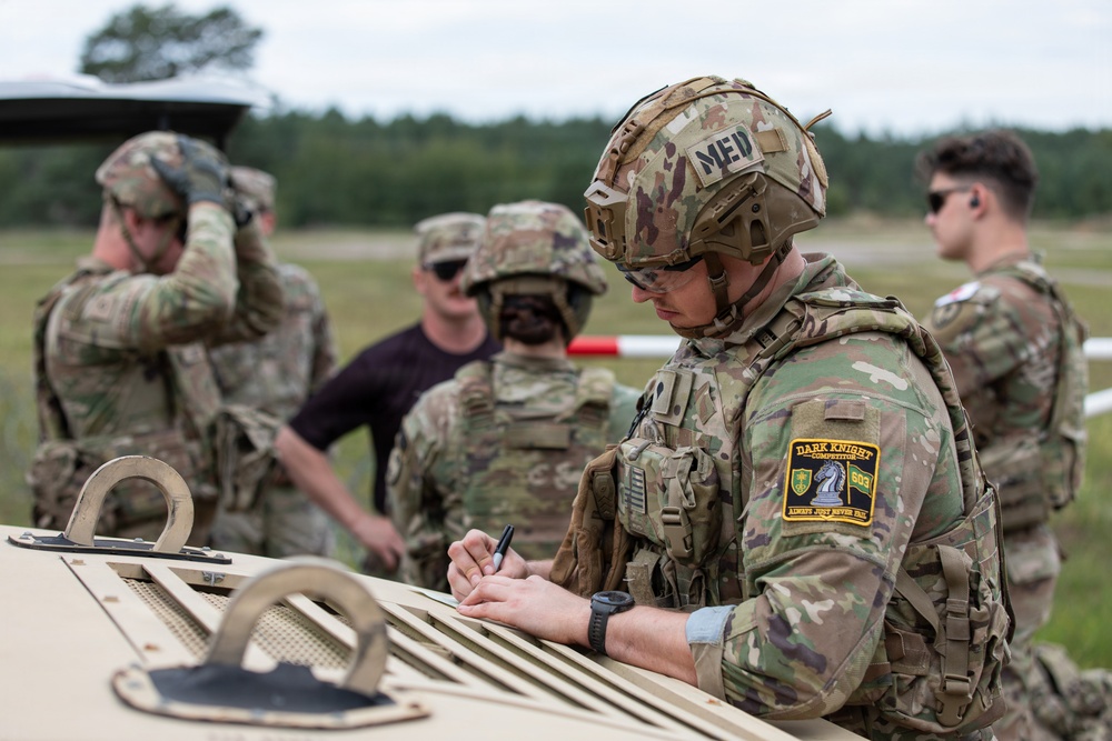 A group of military personnel in MultiCam/OCP camouflage uniforms are gathered outdoors near a military vehicle, likely during a training exercise or competition. The foreground soldier, identified as a medic ('MED' label on helmet), is writing on a clipboard or document placed on the vehicle's surface. A patch on his vest reads 'Dark Knight Competitor 603' with the motto 'Always Just Never Fail.'