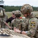 A group of military personnel in MultiCam/OCP camouflage uniforms are gathered outdoors near a military vehicle, likely during a training exercise or competition. The foreground soldier, identified as a medic ('MED' label on helmet), is writing on a clipboard or document placed on the vehicle's surface. A patch on his vest reads 'Dark Knight Competitor 603' with the motto 'Always Just Never Fail.'