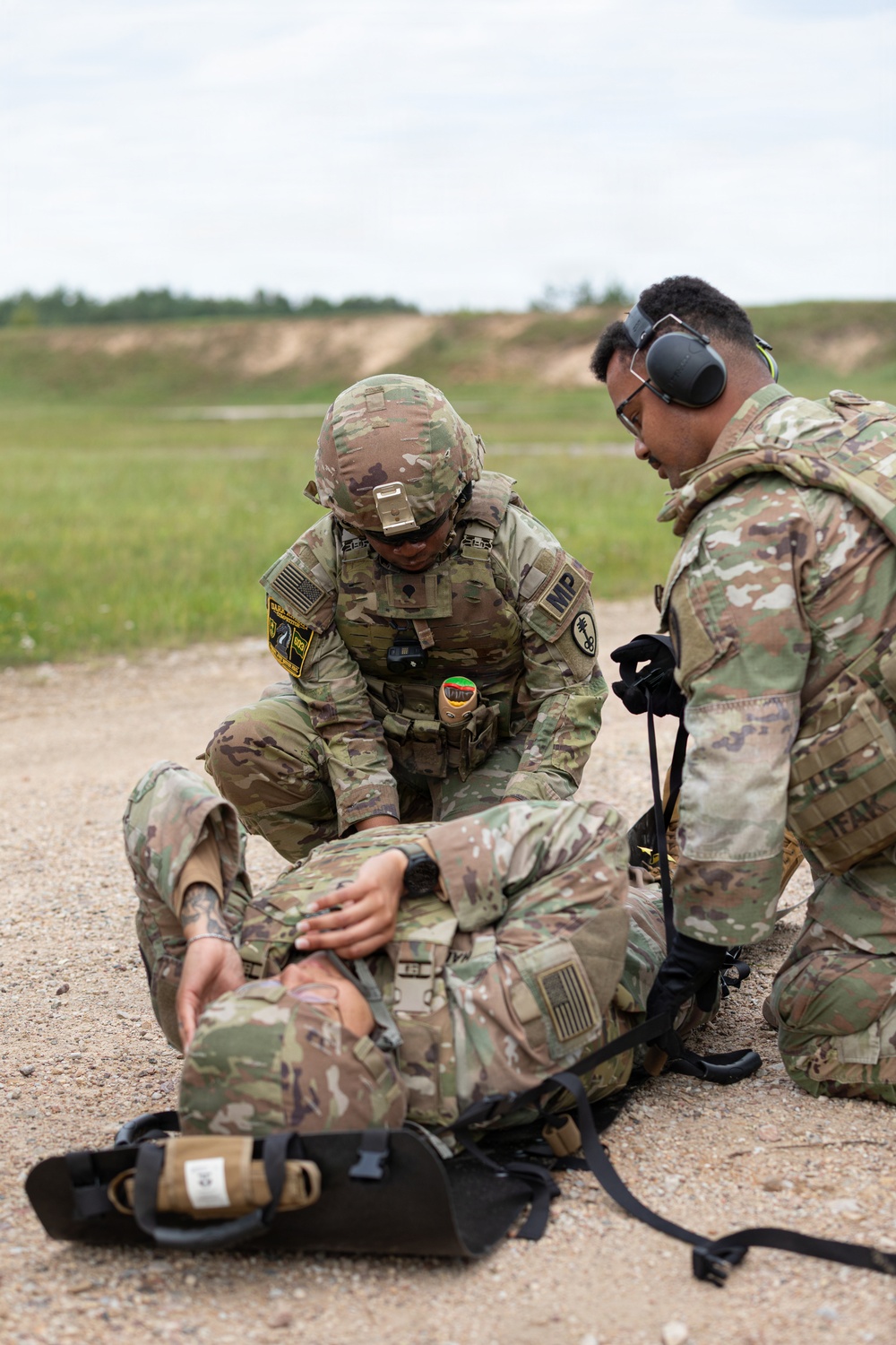Two U.S. Army soldiers in Operational Camouflage Pattern (OCP) uniforms are conducting a tactical combat casualty care (TCCC) exercise on a gravel surface at what appears to be a training range. One soldier wearing a combat helmet with an 'MP' (Military Police) patch is treating a simulated casualty on the ground, while a second soldier wearing hearing protection and eye protection assists. Medical equipment is visible on the ground nearby.