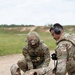 Two U.S. Army soldiers in Operational Camouflage Pattern (OCP) uniforms are conducting a tactical combat casualty care (TCCC) exercise on a gravel surface at what appears to be a training range. One soldier wearing a combat helmet with an 'MP' (Military Police) patch is treating a simulated casualty on the ground, while a second soldier wearing hearing protection and eye protection assists. Medical equipment is visible on the ground nearby.