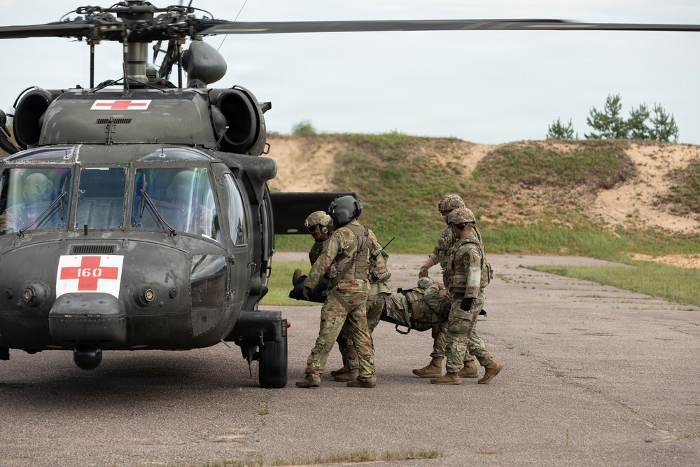 Military personnel in camouflage uniforms are loading a casualty on a litter into a medical evacuation helicopter. The helicopter is a UH-60 Black Hawk marked with red cross insignia and the number 160, indicating it is configured for MEDEVAC operations. The scene appears to be a training exercise or actual casualty evacuation on a paved landing area.