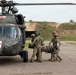 Military personnel in camouflage uniforms are loading a casualty on a litter into a medical evacuation helicopter. The helicopter is a UH-60 Black Hawk marked with red cross insignia and the number 160, indicating it is configured for MEDEVAC operations. The scene appears to be a training exercise or actual casualty evacuation on a paved landing area.