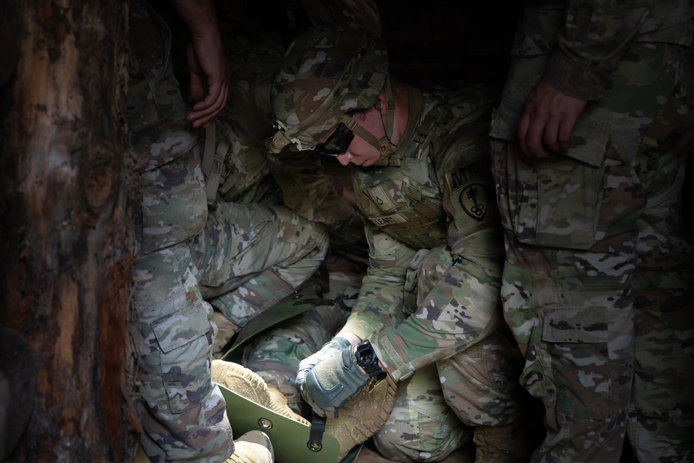 The image shows military personnel in camouflage uniforms (OCP/MultiCam pattern) in what appears to be a confined space or trench. A central soldier wearing tactical goggles and gloves is crouched down, appearing to examine or work with a green military clipboard or document holder. The scene suggests a training exercise or field operation.