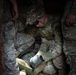 The image shows military personnel in camouflage uniforms (OCP/MultiCam pattern) in what appears to be a confined space or trench. A central soldier wearing tactical goggles and gloves is crouched down, appearing to examine or work with a green military clipboard or document holder. The scene suggests a training exercise or field operation.