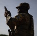 A soldier in multicam/OCP camouflage uniform is photographed from behind, holding a pistol in a raised position consistent with a weapons inspection or clearing drill. A unit patch on the left sleeve reads 'Dark Knight' with what appears to be a chess knight insignia and the number 663. The image is backlit, creating a silhouette-like effect against a partly cloudy sky.