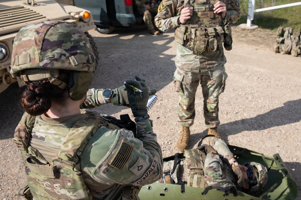 A U.S. Army soldier named Gonzalez is kneeling and administering or preparing a medical treatment on a simulated casualty lying on a litter/stretcher during what appears to be a combat casualty care training exercise. A second standing soldier with the name tag 'LINDGREN' observes the procedure, likely acting as an evaluator or instructor. The setting appears to be an outdoor training area with military vehicles present.