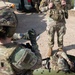 A U.S. Army soldier named Gonzalez is kneeling and administering or preparing a medical treatment on a simulated casualty lying on a litter/stretcher during what appears to be a combat casualty care training exercise. A second standing soldier with the name tag 'LINDGREN' observes the procedure, likely acting as an evaluator or instructor. The setting appears to be an outdoor training area with military vehicles present.