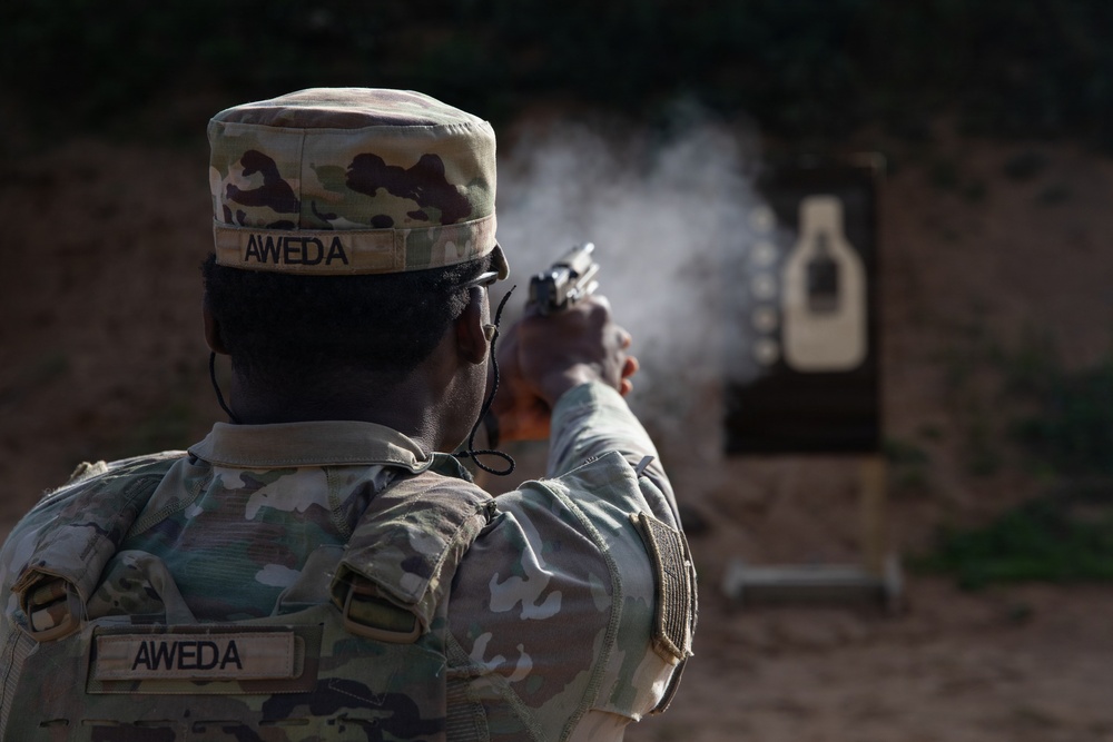 A military service member with the name tape 'AWEDA' is firing a pistol at a silhouette target on a shooting range. The image captures the moment of discharge, with visible muzzle smoke rising from the weapon. The soldier is wearing MultiCam/OCP camouflage uniform and patrol cap.
