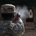 A military service member with the name tape 'AWEDA' is firing a pistol at a silhouette target on a shooting range. The image captures the moment of discharge, with visible muzzle smoke rising from the weapon. The soldier is wearing MultiCam/OCP camouflage uniform and patrol cap.
