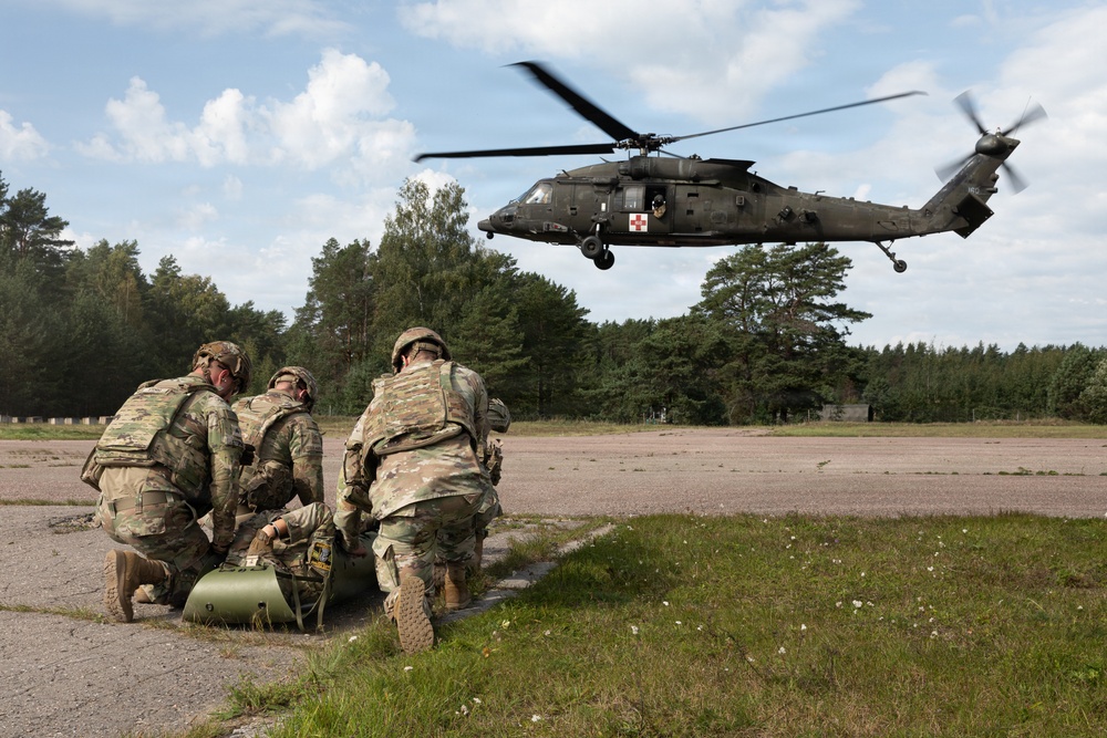 Three soldiers in camouflage uniforms kneel on a paved surface while tending to a casualty on a litter, as a military medevac helicopter (UH-60 Black Hawk) hovers nearby. The helicopter displays a red cross emblem, indicating it is configured for medical evacuation. This appears to be a MEDEVAC training exercise or real casualty evacuation operation.