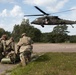 Three soldiers in camouflage uniforms kneel on a paved surface while tending to a casualty on a litter, as a military medevac helicopter (UH-60 Black Hawk) hovers nearby. The helicopter displays a red cross emblem, indicating it is configured for medical evacuation. This appears to be a MEDEVAC training exercise or real casualty evacuation operation.