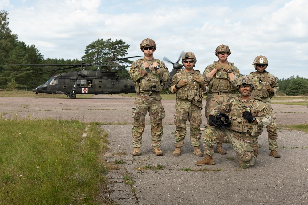 Five military personnel in full combat gear pose for a group photo on a tarmac or landing zone. Behind them is a UH-60 Black Hawk helicopter marked with a red cross, indicating a MEDEVAC (medical evacuation) configuration. The setting appears to be a military training area or forward operating base surrounded by pine trees.