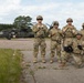 Five military personnel in full combat gear pose for a group photo on a tarmac or landing zone. Behind them is a UH-60 Black Hawk helicopter marked with a red cross, indicating a MEDEVAC (medical evacuation) configuration. The setting appears to be a military training area or forward operating base surrounded by pine trees.