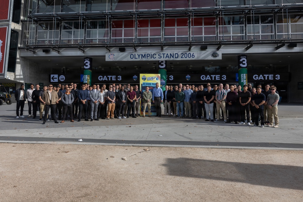 A large group of approximately 60-80 people pose for a group photograph outside Gate 3 of the Olympic Stand 2006 at Melbourne Park. The gathering appears to be associated with a sporting event, as indicated by promotional signage visible in the background referencing a 'Kickoff' game or event. The group consists predominantly of young men dressed in smart-casual attire standing in a wide formation across the stadium entrance.