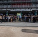 A large group of approximately 60-80 people pose for a group photograph outside Gate 3 of the Olympic Stand 2006 at Melbourne Park. The gathering appears to be associated with a sporting event, as indicated by promotional signage visible in the background referencing a 'Kickoff' game or event. The group consists predominantly of young men dressed in smart-casual attire standing in a wide formation across the stadium entrance.