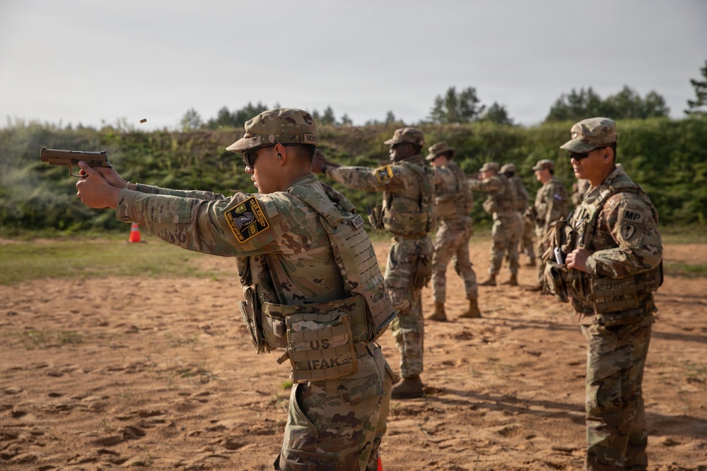 A group of U.S. Army soldiers in OCP (Operational Camouflage Pattern) uniforms are conducting pistol qualification or training on an outdoor firing range. The foreground soldier is actively firing a semi-automatic pistol, with a spent casing visibly ejecting from the weapon. An MP (Military Police) soldier observes from the right side, likely serving as a range safety officer or instructor.