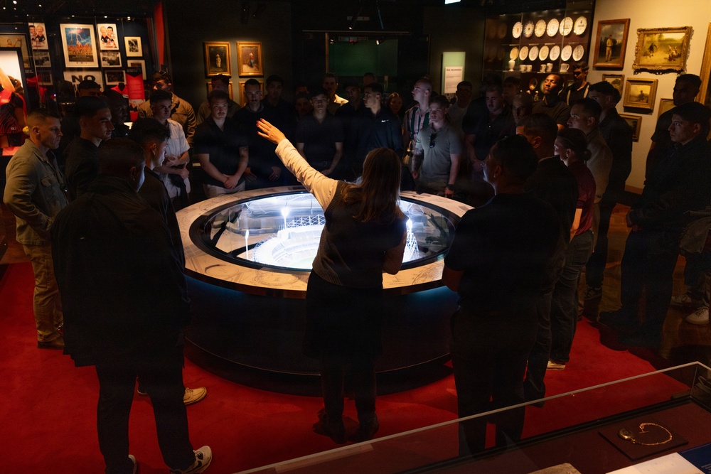 A museum guide or docent is presenting to a large group of visitors gathered around a circular illuminated display case containing what appears to be an architectural scale model. The presenter has one hand raised, actively explaining the exhibit to the attentive crowd. The setting is a dimly lit museum gallery with framed artwork, portraits, and decorative plates visible on the surrounding walls.