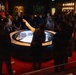 A museum guide or docent is presenting to a large group of visitors gathered around a circular illuminated display case containing what appears to be an architectural scale model. The presenter has one hand raised, actively explaining the exhibit to the attentive crowd. The setting is a dimly lit museum gallery with framed artwork, portraits, and decorative plates visible on the surrounding walls.