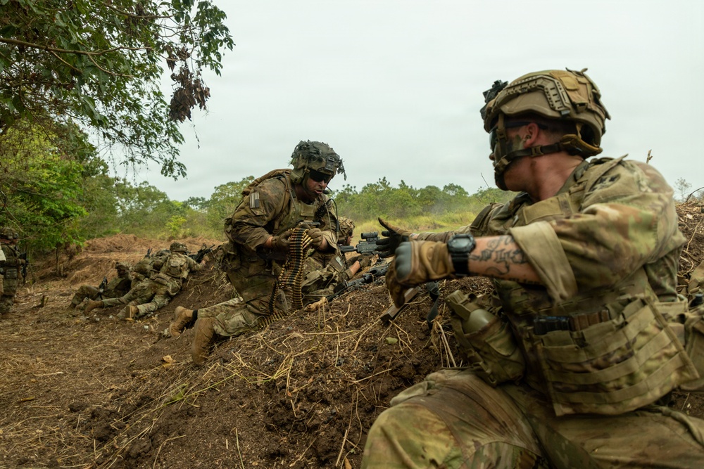 The image shows multiple soldiers in full combat gear positioned in a dirt trench or fighting position in a tropical or subtropical environment. Two soldiers are prominently visible in the foreground, with one handling a belt-fed ammunition chain and another aiming a rifle. Additional soldiers are visible in prone positions in the background.