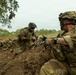 The image shows multiple soldiers in full combat gear positioned in a dirt trench or fighting position in a tropical or subtropical environment. Two soldiers are prominently visible in the foreground, with one handling a belt-fed ammunition chain and another aiming a rifle. Additional soldiers are visible in prone positions in the background.