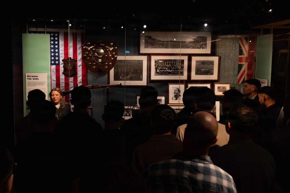 A museum guide or curator is presenting to a group of visitors in front of a dimly lit exhibition display case. The exhibit appears to be titled 'Between the Wars' and features historical football/soccer memorabilia including flags, trophies, and archival photographs. The crowd is gathered closely, listening to the presenter who stands to the left of the display.