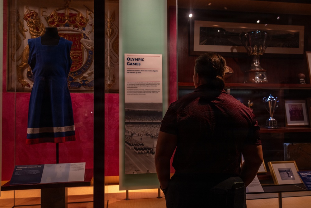 A visitor stands in a museum exhibition hall reading an 'Olympic Games' display panel, which references Melbourne and the MCG. To the left, a blue dress is displayed on a mannequin inside a glass case, with a royal coat of arms visible in the background. The exhibit appears to relate to Melbourne's history, including the 1956 Olympic Games.