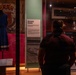A visitor stands in a museum exhibition hall reading an 'Olympic Games' display panel, which references Melbourne and the MCG. To the left, a blue dress is displayed on a mannequin inside a glass case, with a royal coat of arms visible in the background. The exhibit appears to relate to Melbourne's history, including the 1956 Olympic Games.