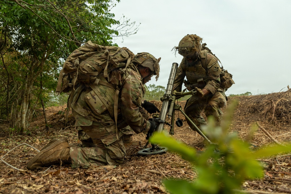 Two soldiers in full combat gear and camouflage uniforms are setting up what appears to be an M224 or M252 mortar system in a field environment with vegetation in the background. Both soldiers are wearing MultiCam/OCP pattern uniforms with tactical vests, helmets with camouflage covers, and large rucksacks. The image appears to depict a military training exercise or field operation.