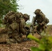 Two soldiers in full combat gear and camouflage uniforms are setting up what appears to be an M224 or M252 mortar system in a field environment with vegetation in the background. Both soldiers are wearing MultiCam/OCP pattern uniforms with tactical vests, helmets with camouflage covers, and large rucksacks. The image appears to depict a military training exercise or field operation.