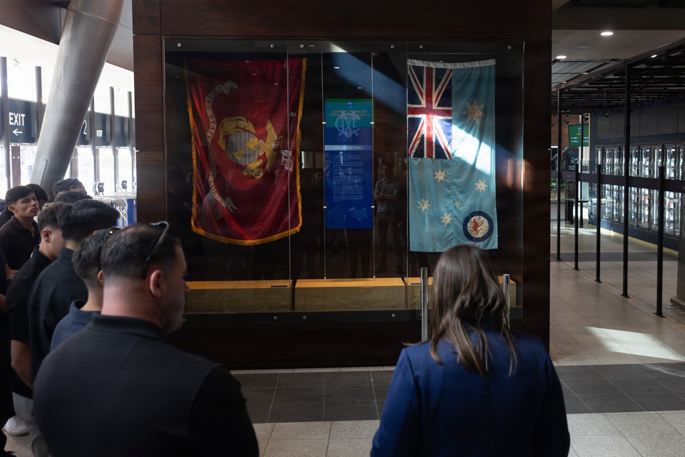 A group of people are viewing a military flag display case in what appears to be a public building or museum. The display features two prominent flags: a United States Marine Corps flag on the left and what appears to be a Royal Australian Air Force (RAAF) Ensign on the right. Visitors, including a woman in a blue blazer and several young men in dark clothing, are observing the exhibit.