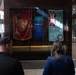 A group of people are viewing a military flag display case in what appears to be a public building or museum. The display features two prominent flags: a United States Marine Corps flag on the left and what appears to be a Royal Australian Air Force (RAAF) Ensign on the right. Visitors, including a woman in a blue blazer and several young men in dark clothing, are observing the exhibit.