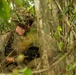 A soldier in full combat gear is positioned within dense jungle vegetation, partially concealed by foliage. The individual is equipped with a belt-fed machine gun and wearing a camouflaged helmet with leaf cover, suggesting a reconnaissance or ambush posture. The setting appears to be a tropical forest environment, consistent with a military training exercise or operational deployment.