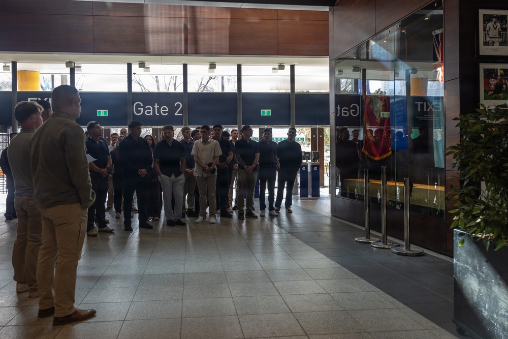 A group of approximately 20-25 people, predominantly young males, are gathered in what appears to be a stadium or arena lobby near Gate 2. Two individuals on the left side appear to be addressing or presenting to the group. The setting includes display cases, framed photographs on the wall, and ceremonial flags, suggesting an official tour or orientation event.