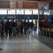 A group of approximately 20-25 people, predominantly young males, are gathered in what appears to be a stadium or arena lobby near Gate 2. Two individuals on the left side appear to be addressing or presenting to the group. The setting includes display cases, framed photographs on the wall, and ceremonial flags, suggesting an official tour or orientation event.