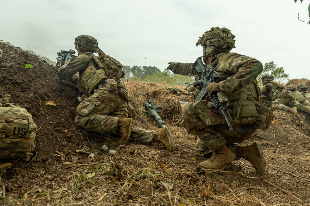 The image shows multiple soldiers in full combat gear positioned along a dirt berm or earthen fortification. The soldiers are wearing MultiCam (OCP) camouflage uniforms with tactical vests and helmets, and are armed with assault rifles. One soldier is pointing forward while others appear to be in firing or observation positions.