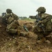 The image shows multiple soldiers in full combat gear positioned along a dirt berm or earthen fortification. The soldiers are wearing MultiCam (OCP) camouflage uniforms with tactical vests and helmets, and are armed with assault rifles. One soldier is pointing forward while others appear to be in firing or observation positions.