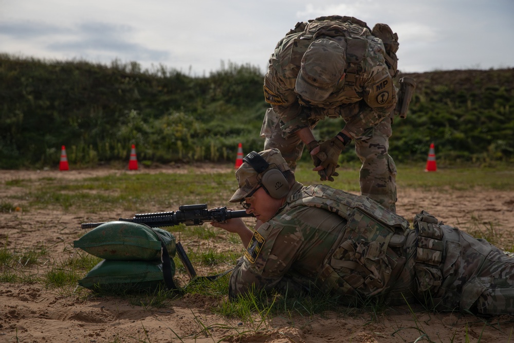 Two military personnel in MultiCam/OCP camouflage uniforms are engaged in a marksmanship training exercise at an outdoor firing range. One soldier is in the prone position aiming a rifle supported by sandbags, while a standing soldier wearing an 'MP' (Military Police) brassard appears to be providing instruction or oversight. Orange traffic cones are visible in the background marking the range area.