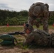 Two military personnel in MultiCam/OCP camouflage uniforms are engaged in a marksmanship training exercise at an outdoor firing range. One soldier is in the prone position aiming a rifle supported by sandbags, while a standing soldier wearing an 'MP' (Military Police) brassard appears to be providing instruction or oversight. Orange traffic cones are visible in the background marking the range area.