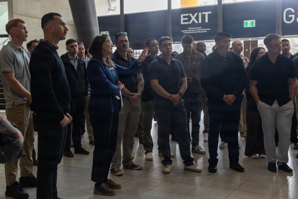A group of approximately 20-30 people are gathered in what appears to be a public venue or stadium concourse area. A woman wearing a blue jacket with a lanyard/badge appears to be leading a tour or briefing, gesturing as she speaks to the assembled group. The setting suggests an organized group visit or orientation event.
