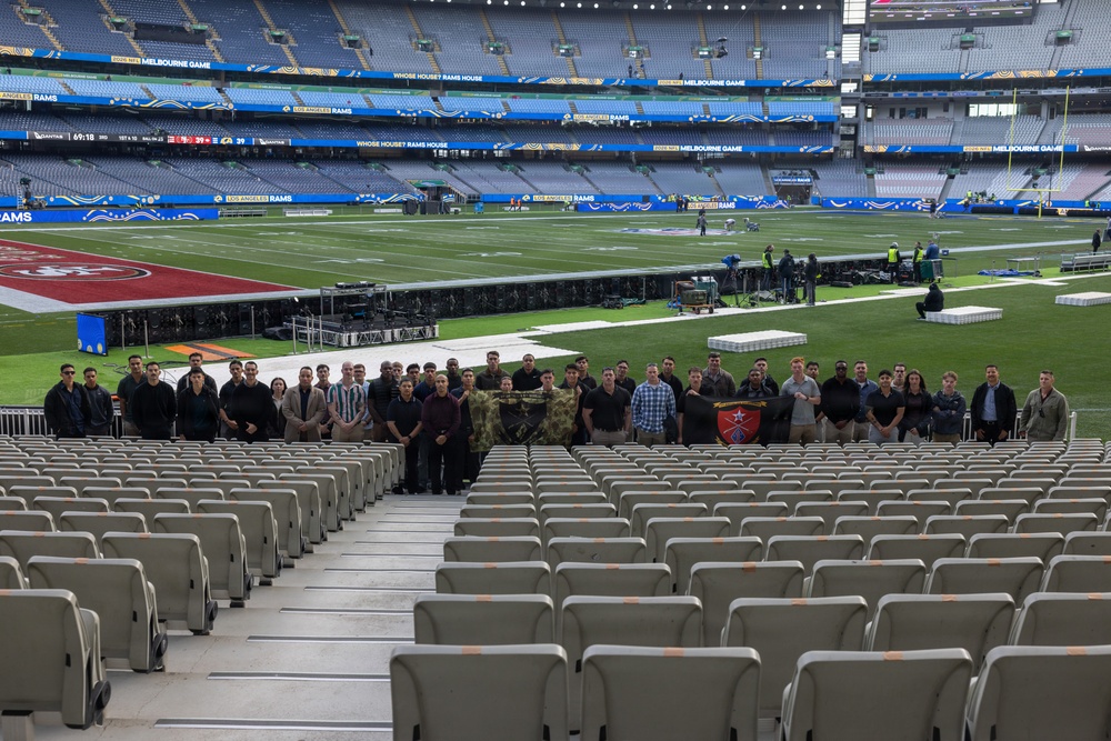 A group of approximately 40 individuals poses for a group photo in the lower seating section of a large stadium configured for an NFL game featuring the Los Angeles Rams, with signage indicating a 'Melbourne Game.' The stadium appears to be the Melbourne Cricket Ground (MCG) in Australia, set up for an NFL international series game. Several people in the group are holding what appears to be a camouflage military flag and a unit insignia banner, suggesting a military group visit.