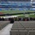 MRF-D Marines tour Melbourne Cricket Ground A group of approximately 40 individuals poses for a group photo in the lower seating section of a large stadium configured for an NFL game featuring the Los Angeles Rams, with signage indicating a 'Melbourne Game.' The stadium appears to be the Melbourne Cricket Ground (MCG) in Australia, set up for an NFL international series game. Several people in the group are holding what appears to be a camouflage military flag and a unit insignia banner, suggesting a military group visit.