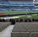 A group of approximately 40 individuals poses for a group photo in the lower seating section of a large stadium configured for an NFL game featuring the Los Angeles Rams, with signage indicating a 'Melbourne Game.' The stadium appears to be the Melbourne Cricket Ground (MCG) in Australia, set up for an NFL international series game. Several people in the group are holding what appears to be a camouflage military flag and a unit insignia banner, suggesting a military group visit.