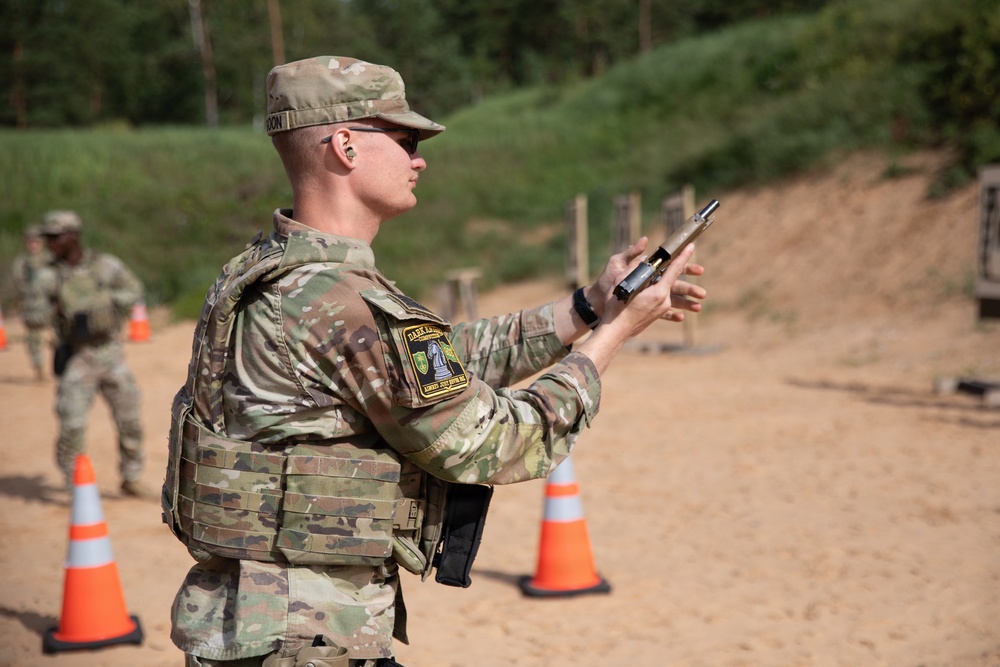 A soldier in MultiCam/OCP camouflage uniform and plate carrier is performing a firearms safety check or clearing procedure with a pistol at what appears to be a military shooting range. The soldier is wearing hearing protection, sunglasses, and a unit patch on the left shoulder. Additional soldiers and orange traffic cones are visible in the background of the sandy range area.