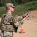A soldier in MultiCam/OCP camouflage uniform and plate carrier is performing a firearms safety check or clearing procedure with a pistol at what appears to be a military shooting range. The soldier is wearing hearing protection, sunglasses, and a unit patch on the left shoulder. Additional soldiers and orange traffic cones are visible in the background of the sandy range area.