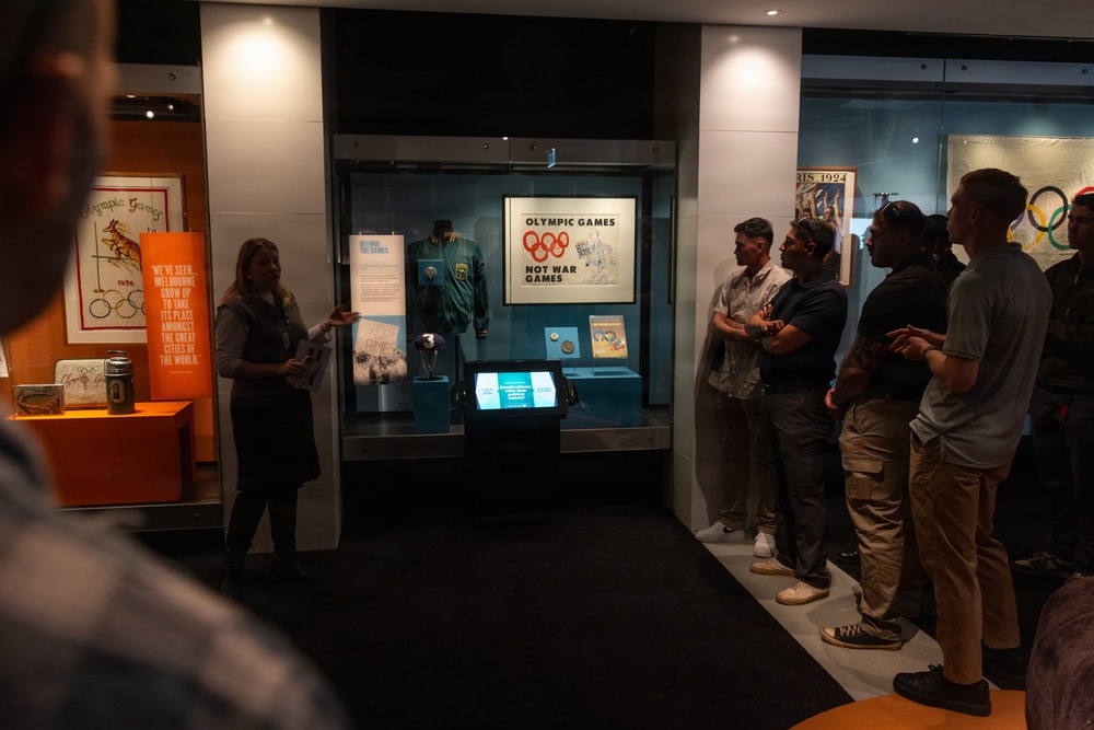 A museum guide is presenting an Olympic Games exhibition to a group of visitors. The display case features artifacts related to the Olympic Games, including a prominent sign reading 'Olympic Games Not War Games.' The exhibit appears to focus on the history of the Olympic Games, with visible references to Melbourne 1956 and Paris 1924.